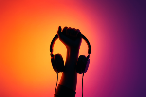 A silhouetted hand holds a pair of headphones against a glowing orange and purple gradient background, symbolizing music streaming and sound.